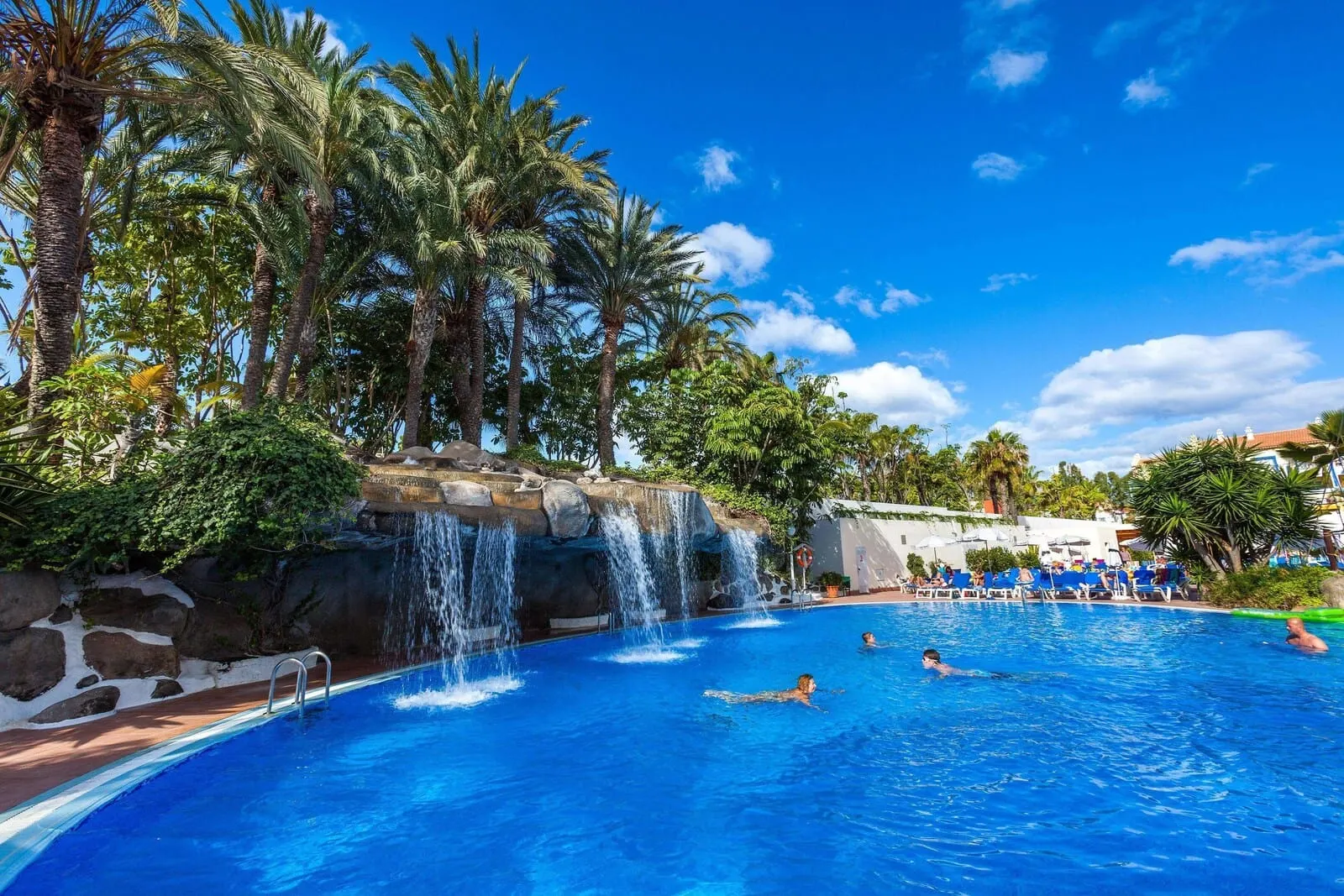 Best Tenerife: A swimming pool with a waterfall feature, surrounded by palm trees and a clear blue sky
