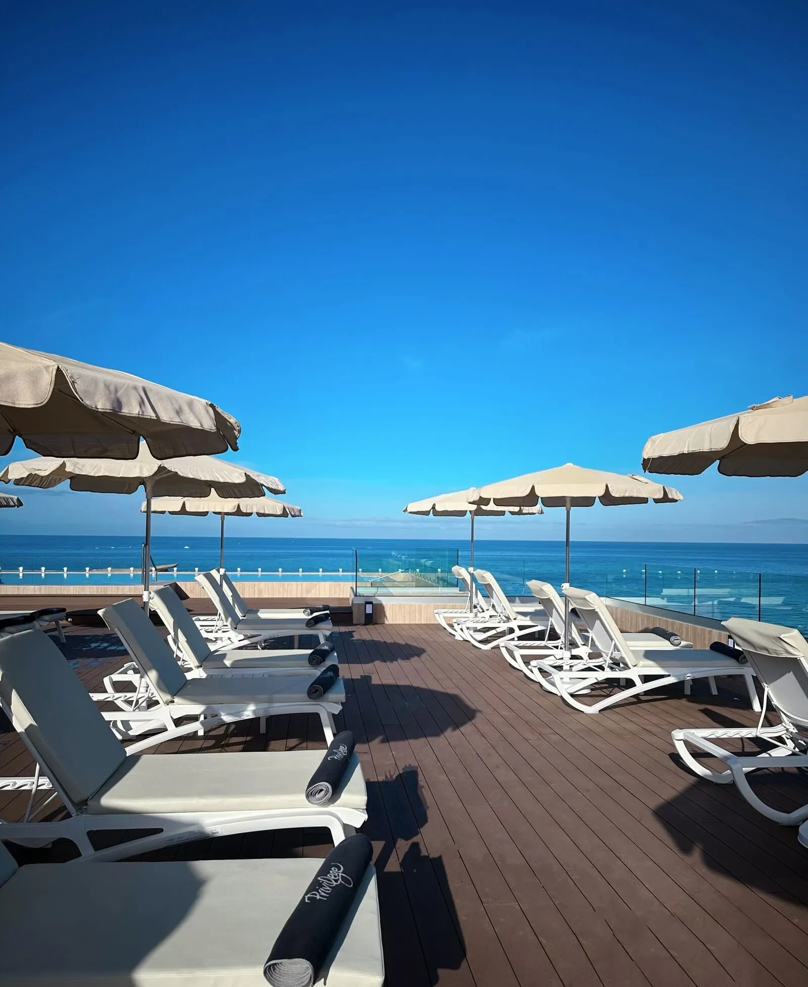 H10 Costa Adeje Palace: A deck with white lounge chairs and beige umbrellas, overlooking a clear blue sea