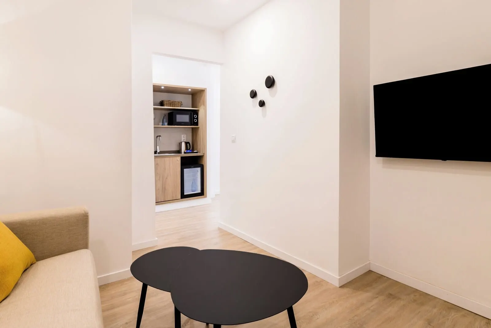Adonis La Suite: A modern living room with a sofa, a black table, and a kitchen area in the background
