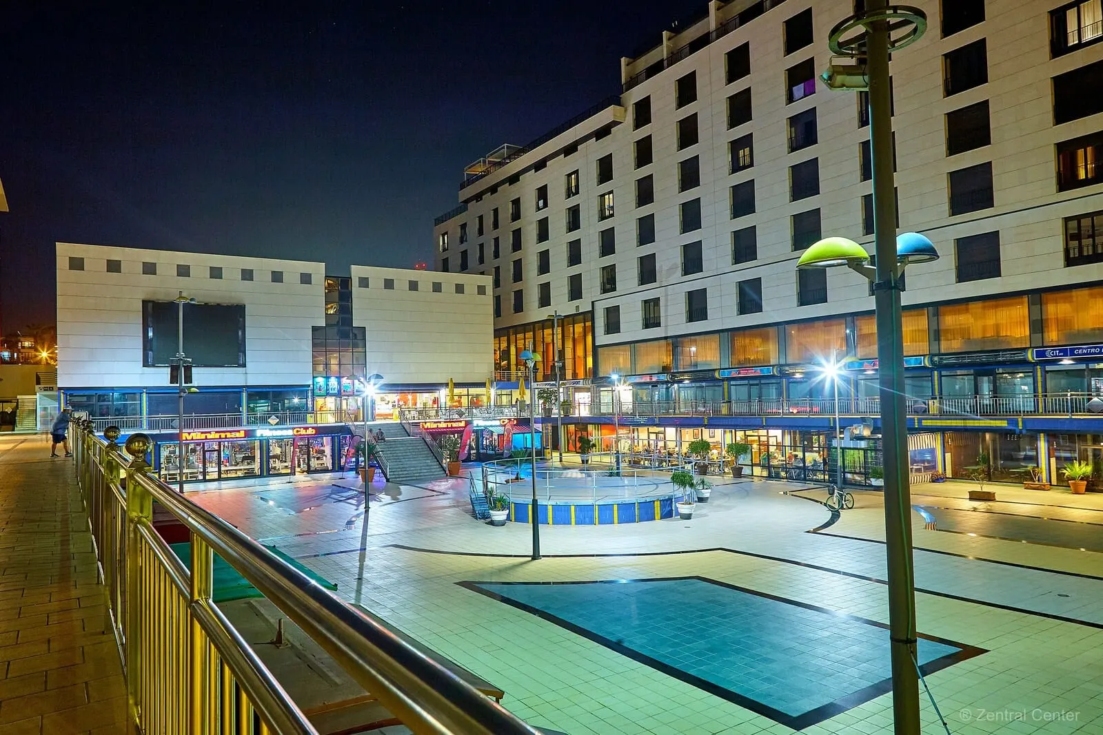 Hotel Zentral Center: A nighttime urban square with a fountain, modern buildings, and a pedestrian walkway