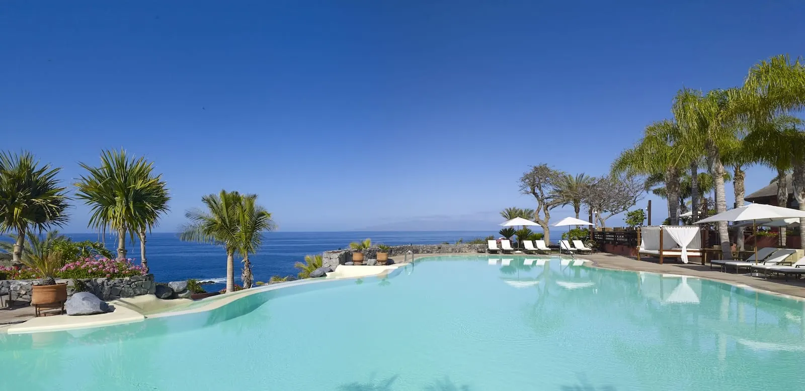 The Ritz-Carlton Tenerife, Abama: A pool with lounge chairs and umbrellas, palm trees, and a view of the ocean