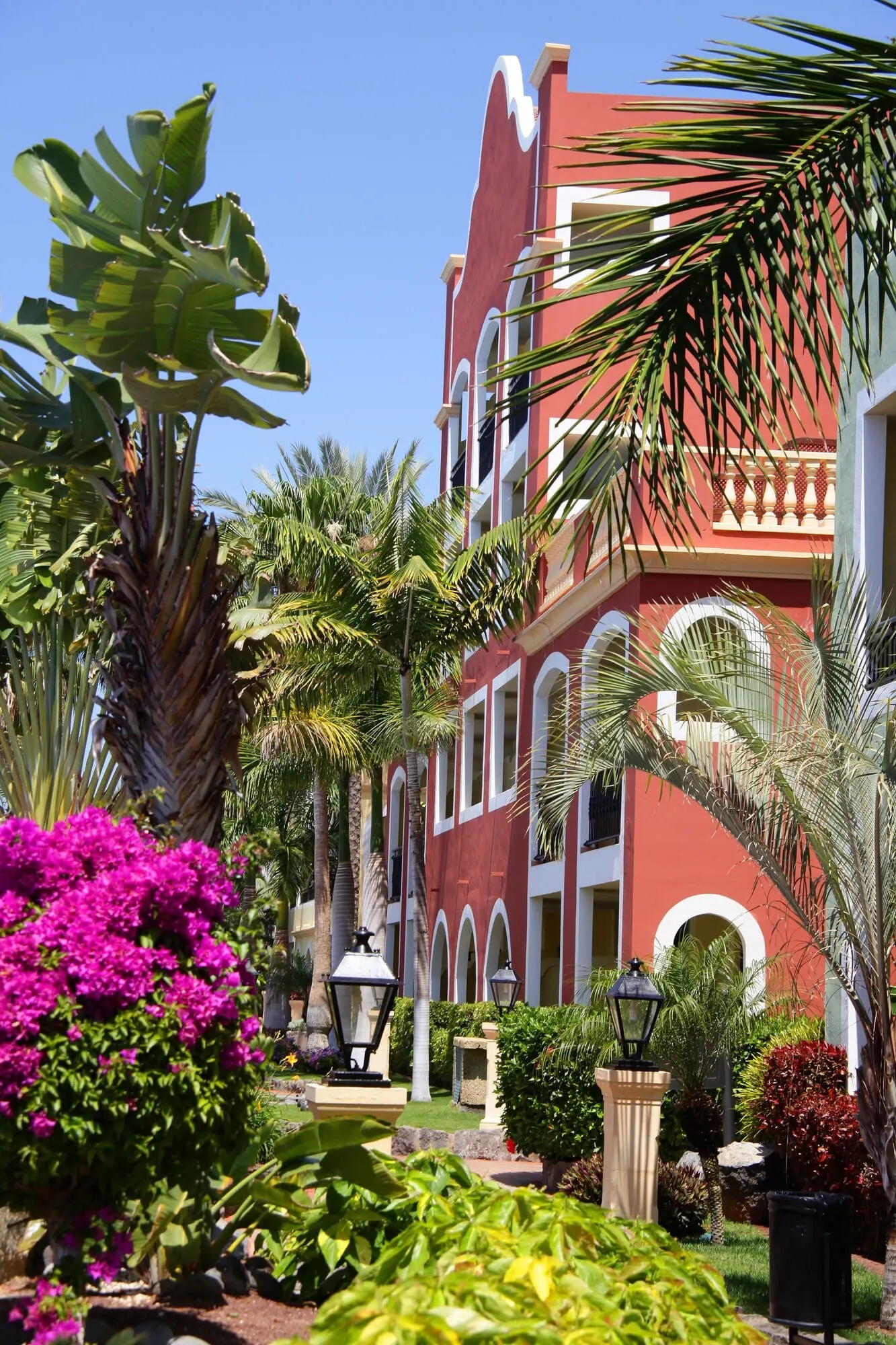 Bahia Principe Explore Costa Adeje: A red building with white arches and a garden with tropical plants and a pink flower