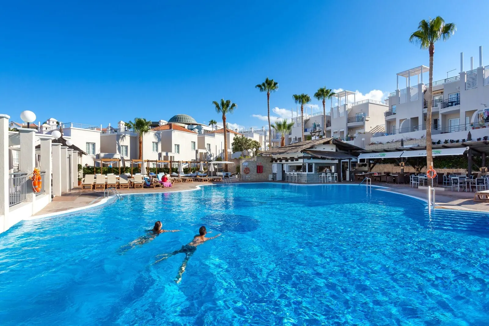Los Olivos Beach Resort: A swimming pool with two people swimming, surrounded by palm trees and modern buildings