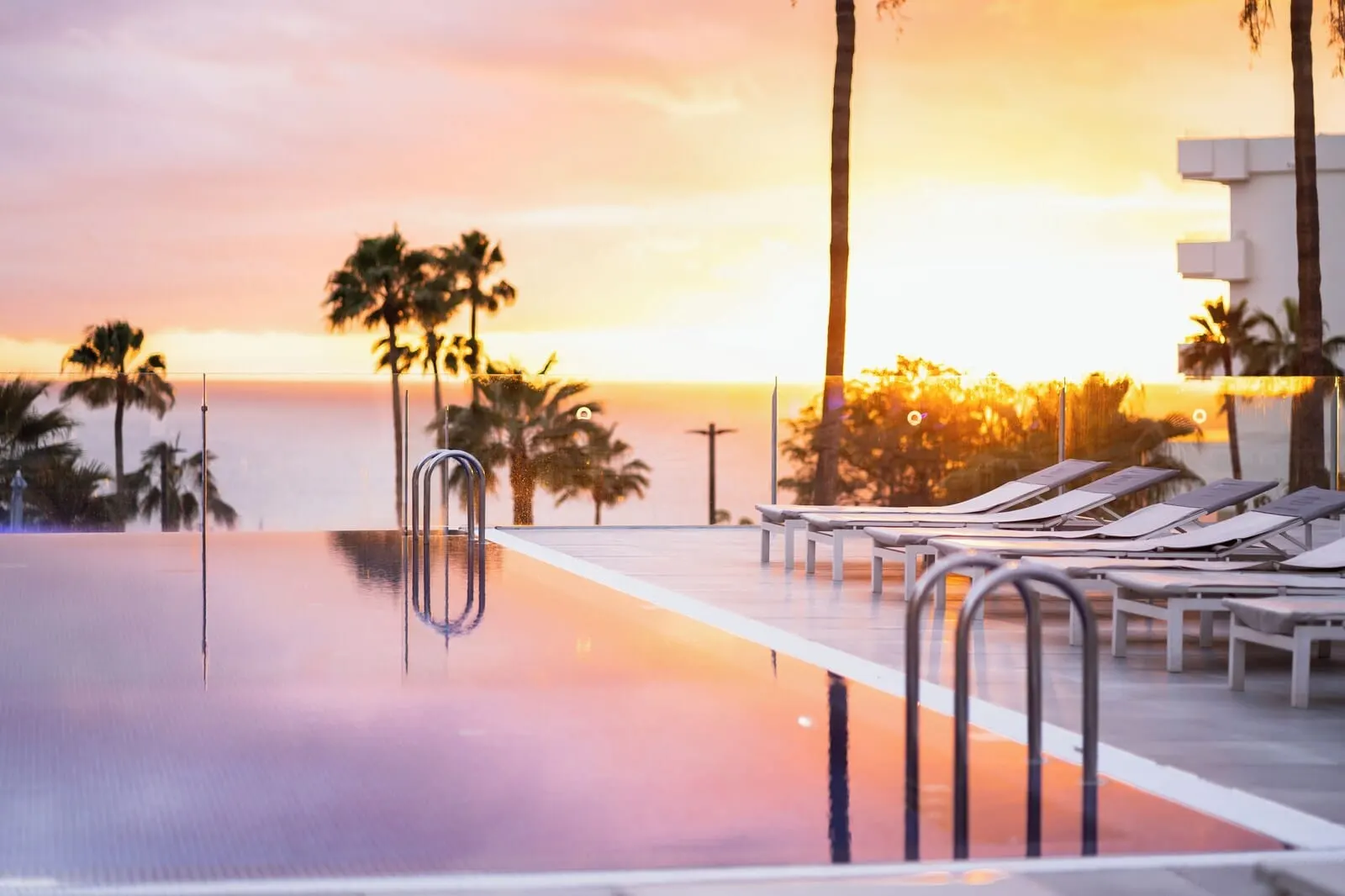 HOVIMA Cool Costa Adeje: An infinity pool with lounge chairs and palm trees, reflecting a sunset