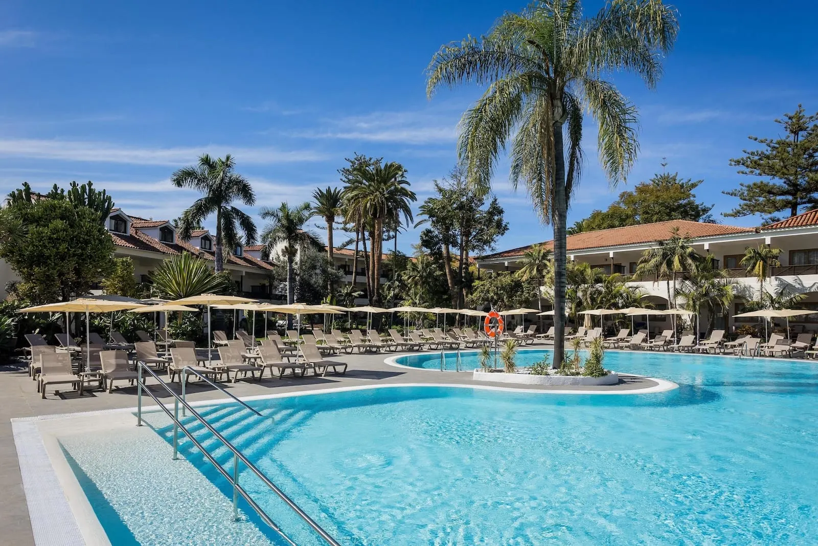 Radisson Resort & Residences Tenerife: A pool area with lounge chairs and umbrellas, surrounded by palm trees and buildings