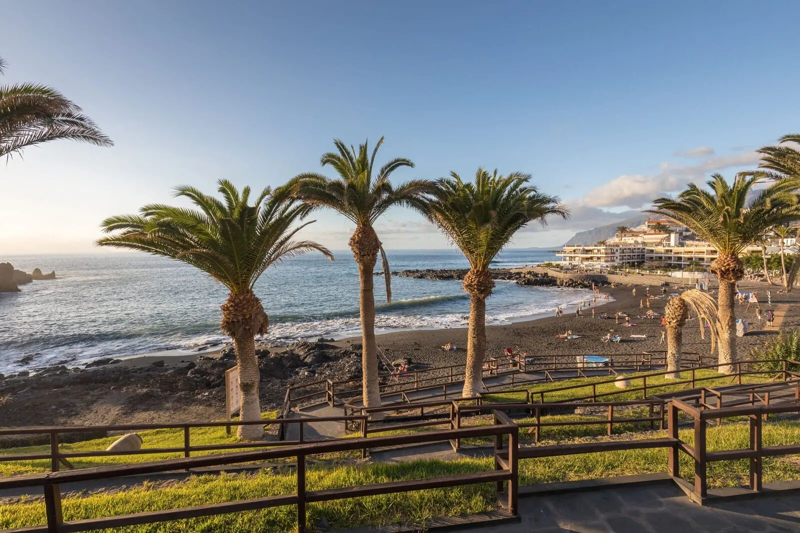 Servatur Isora Suites: A beach with palm trees, a rocky shoreline, and a building complex in the background