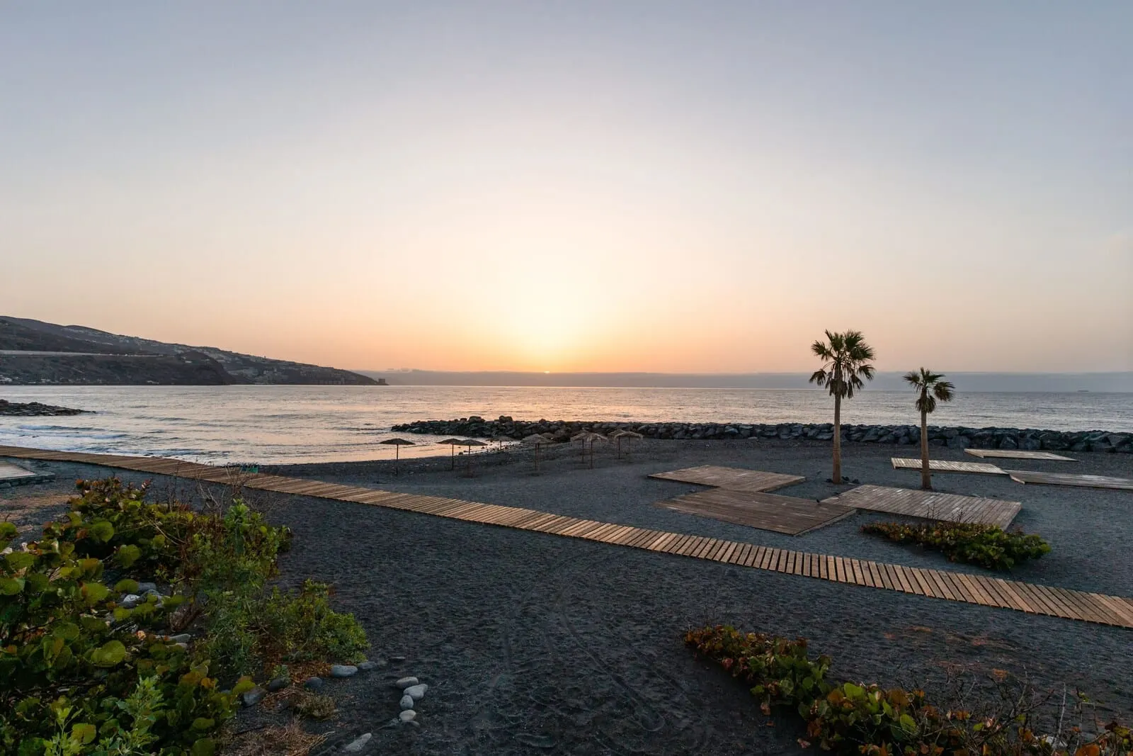 Catalonia Punta del Rey: A beach with wooden walkways, palm trees, and a rocky shoreline at sunset