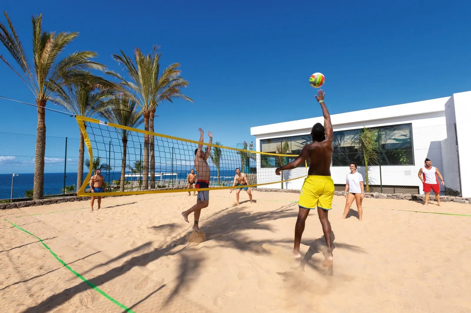Hotel Riu Buenavista: A beach volleyball game on a sandy court with palm trees and a building in