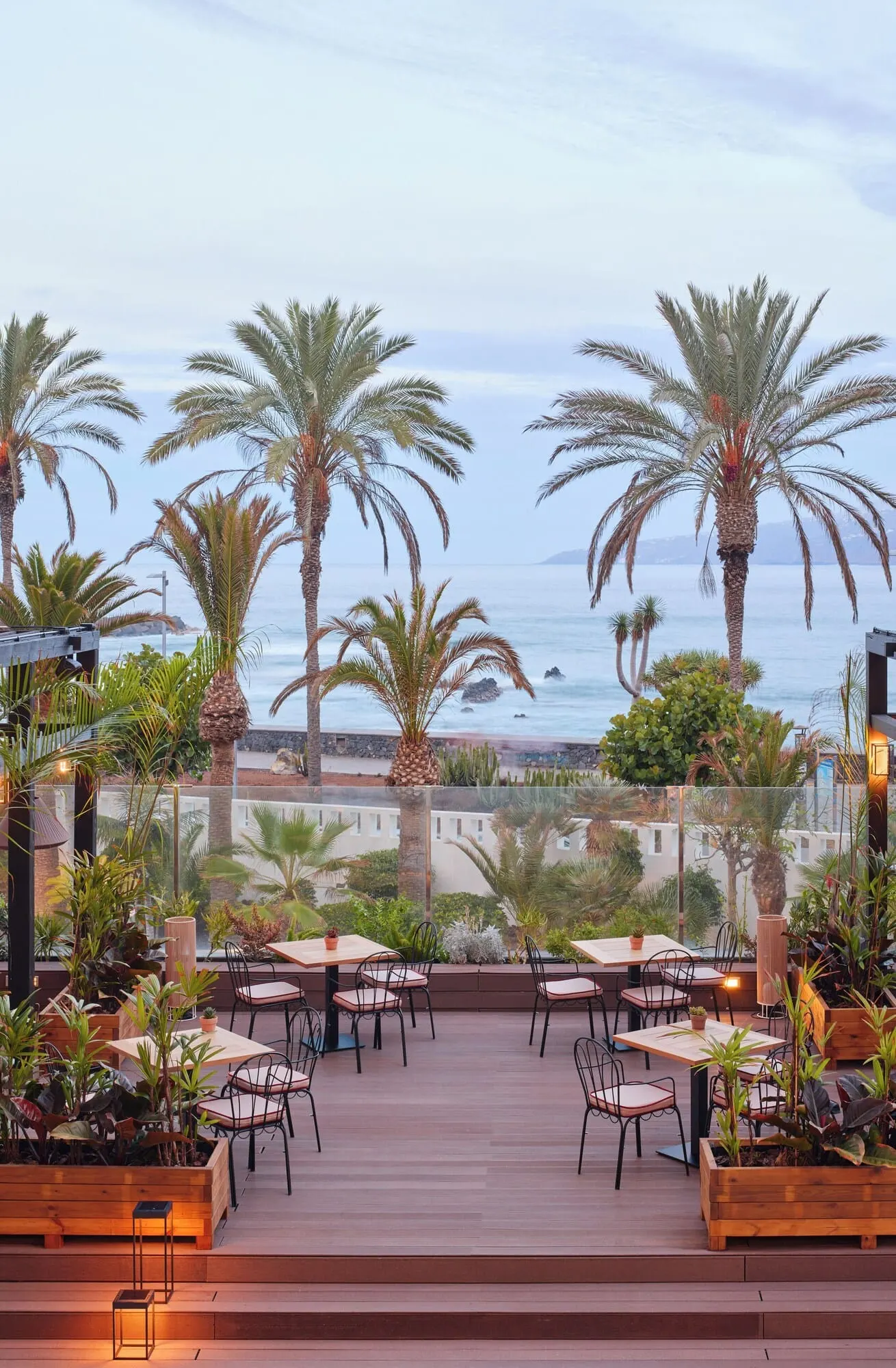 Meliá Costa Atlantis Tenerife: An outdoor patio with wooden tables and chairs, surrounded by palm trees and plants