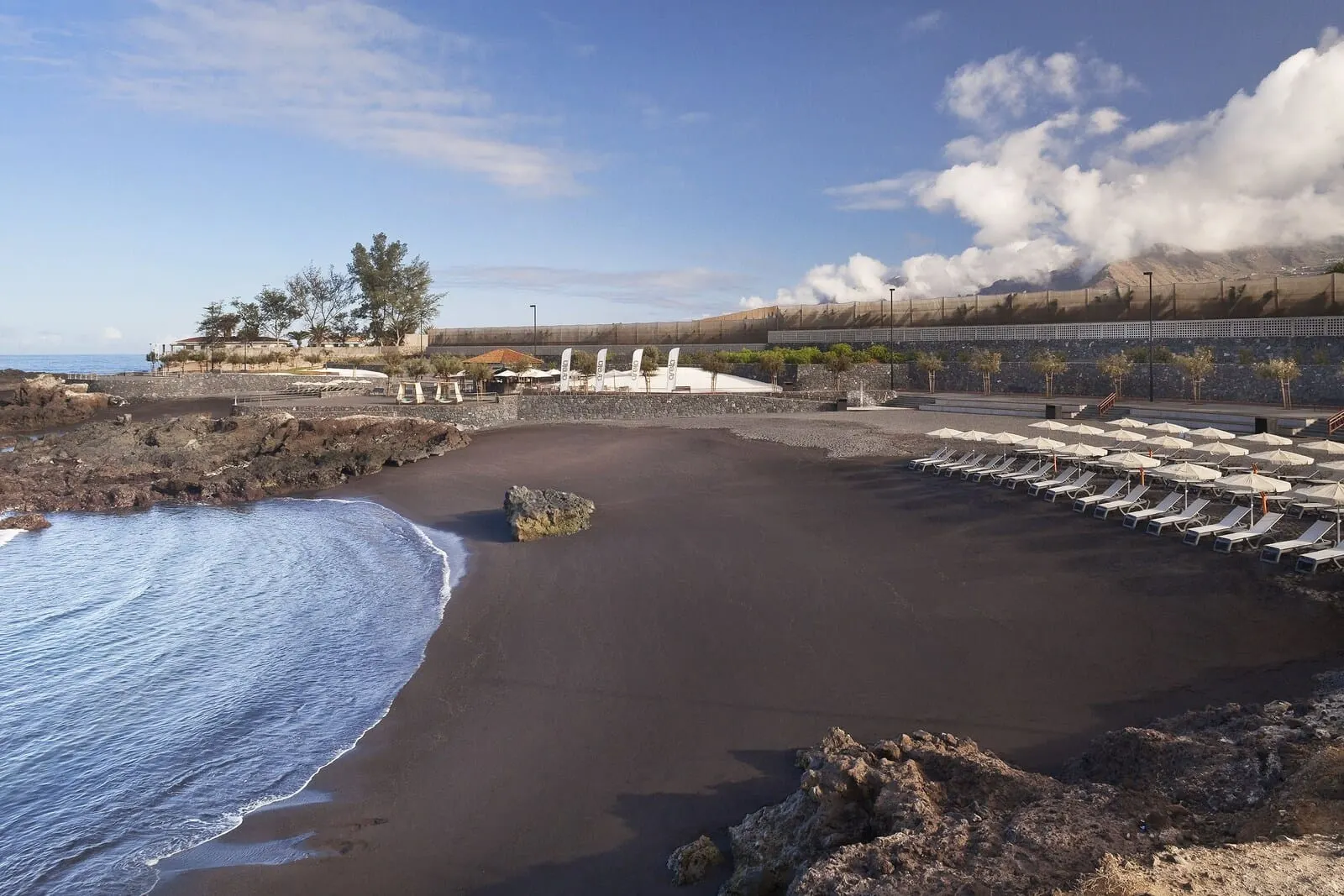 Gran Meliá Palacio de Isora: A beach with a rocky shoreline, a sandy area with lounge chairs, and a distant mountain