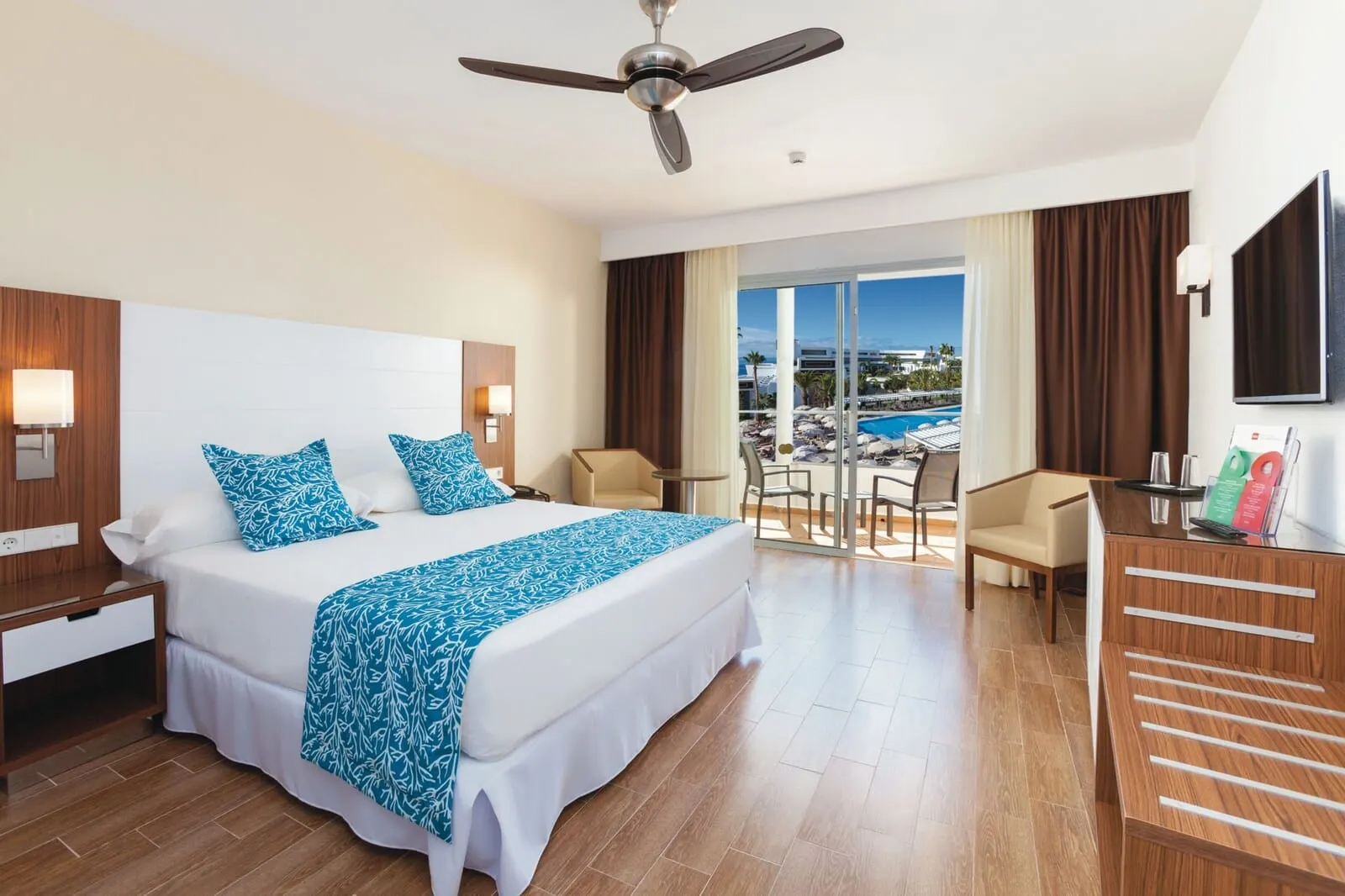 Hotel Riu Arecas: A hotel room with a large bed, a desk, a chair, a TV, and a balcony with a pool view