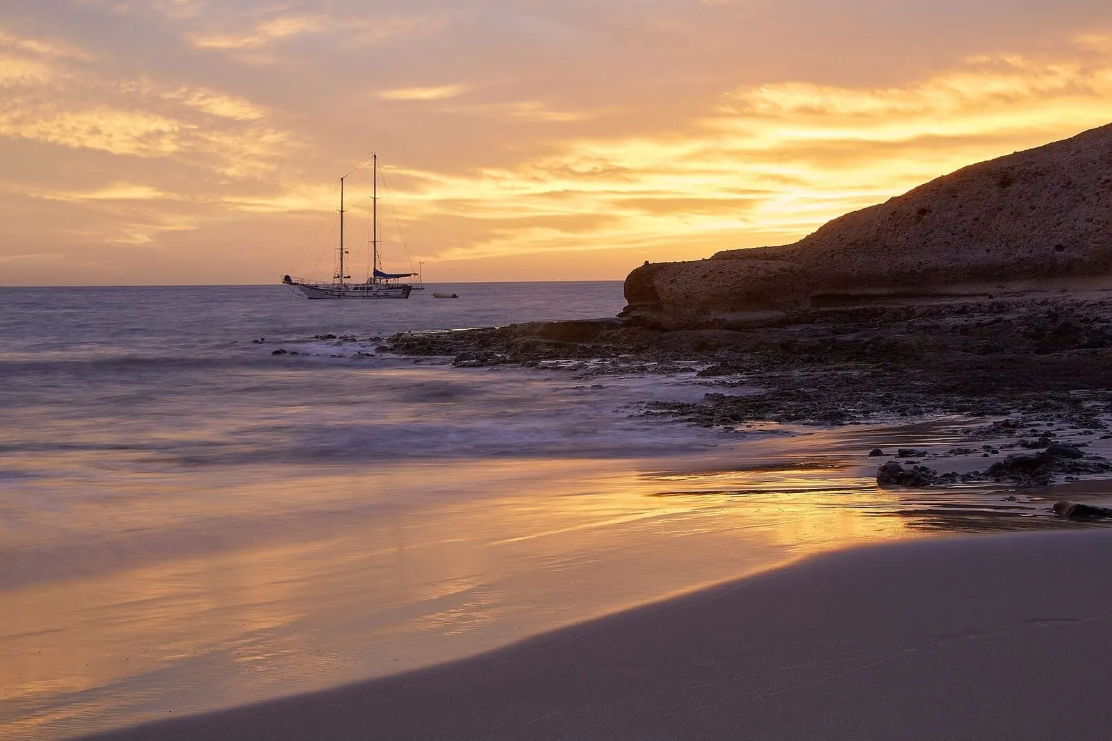 Hotel Suite Villa María: A beach with a sailboat, rocky coastline, and calm waters reflecting sunset