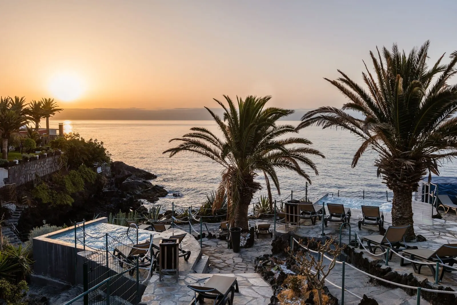Catalonia Punta del Rey: A poolside area with lounge chairs and a hot tub, overlooking a body of water at sunset