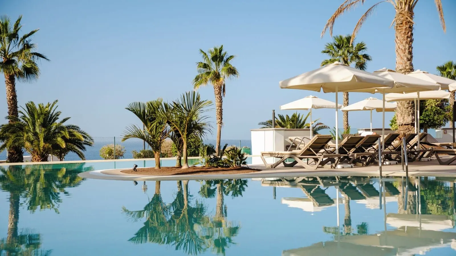 Bahia Principe Escape Tenerife: A pool area with lounge chairs and umbrellas, surrounded by palm trees and a clear sky