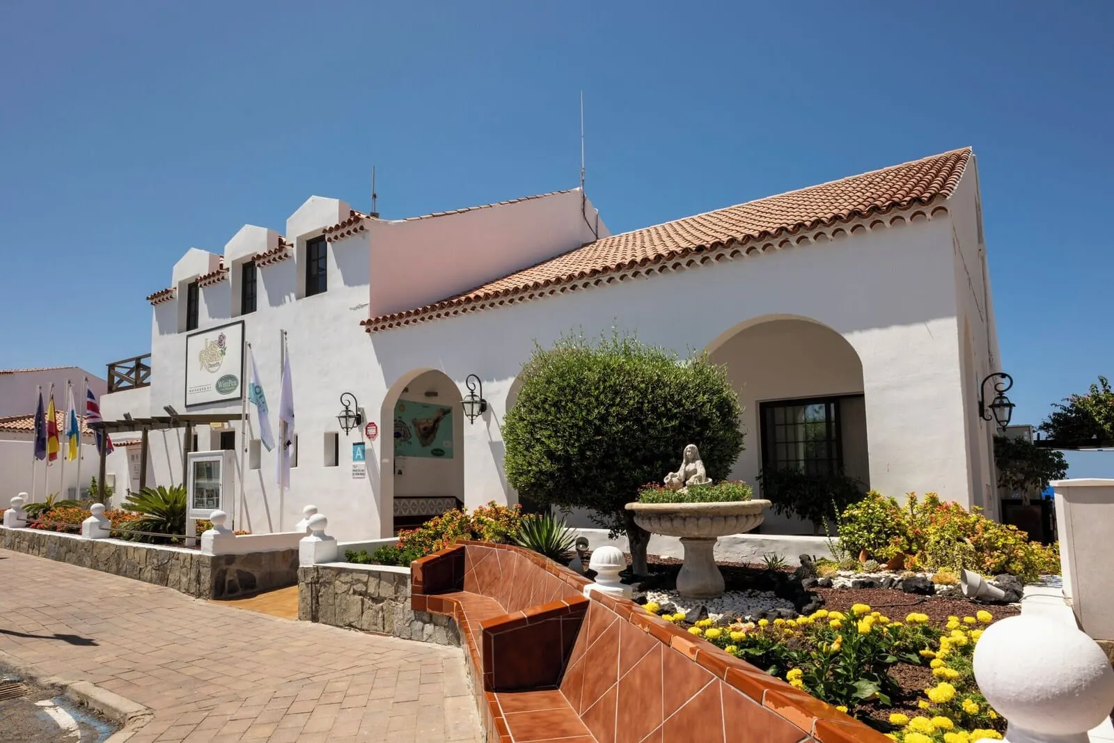 Ona Las Rosas: A white building with a tiled roof, a fountain, and a garden with yellow flowers