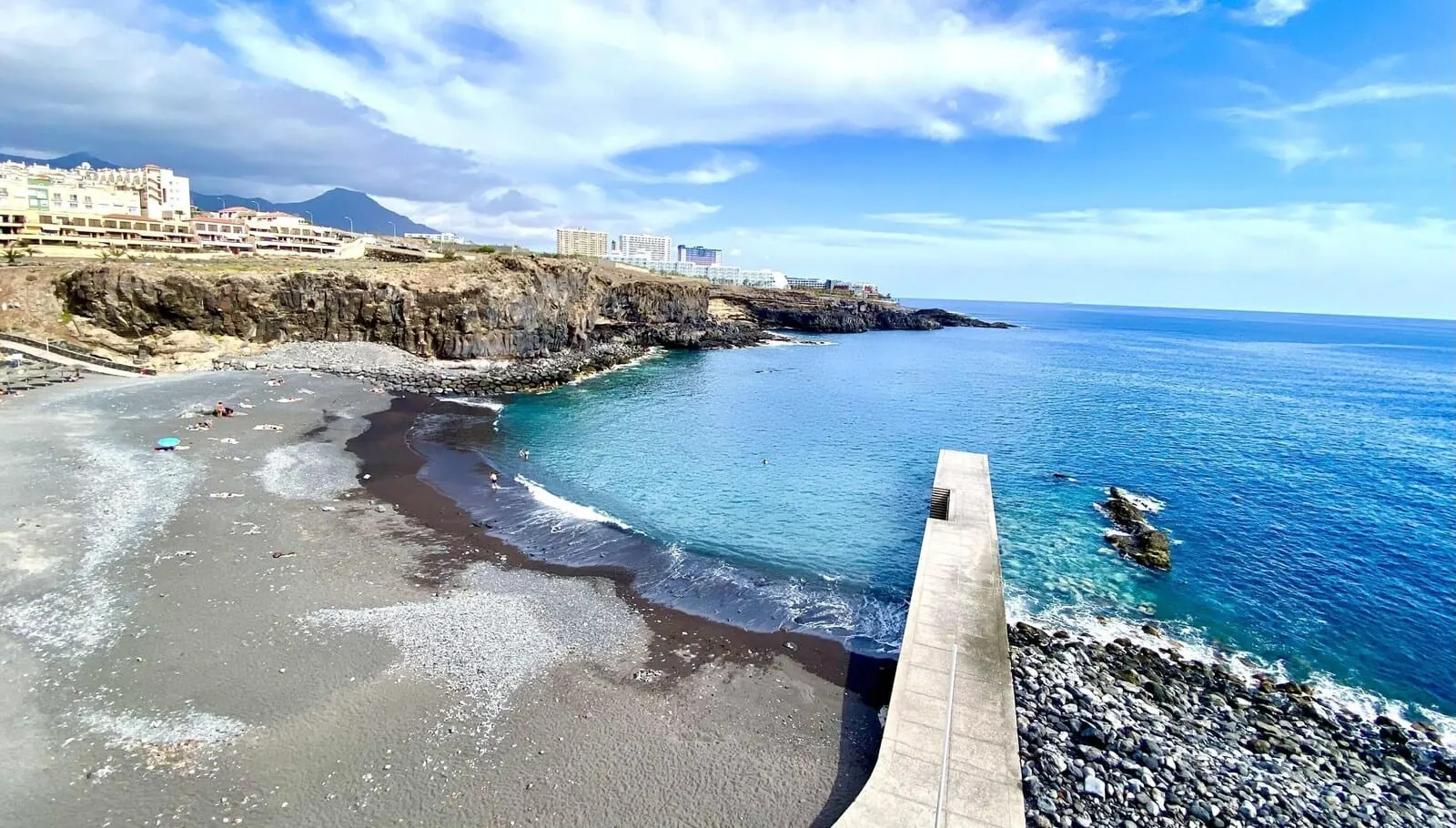 MYND Adeje: A coastal beach with a rocky shoreline, clear blue waters, and a distant mountain