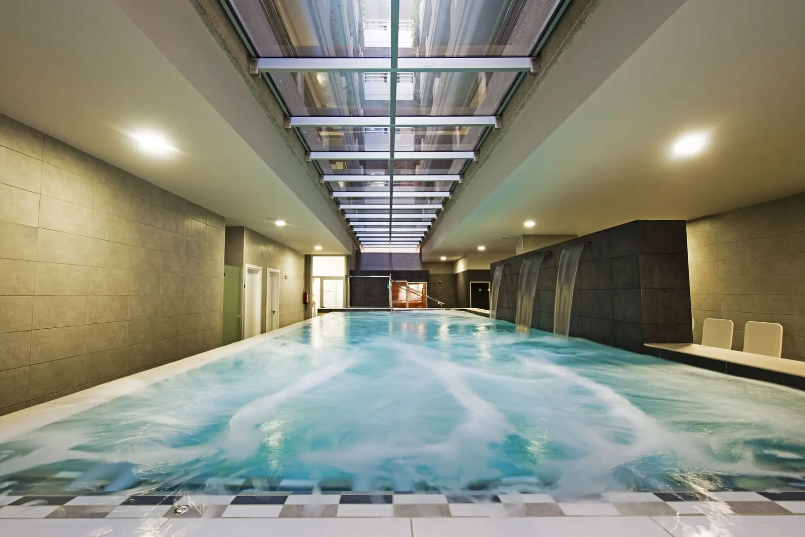 KN Aparthotel Columbus: A large indoor swimming pool with a glass ceiling, tiled walls, and a view of the sky