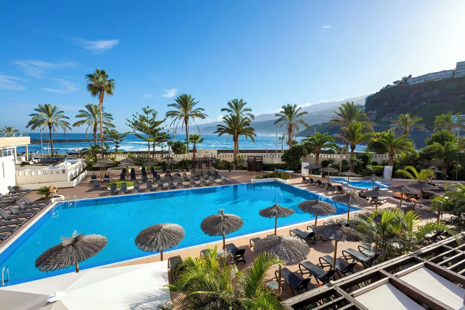 Meliá Costa Atlantis Tenerife: A pool area with lounge chairs and umbrellas, overlooking a beach and ocean