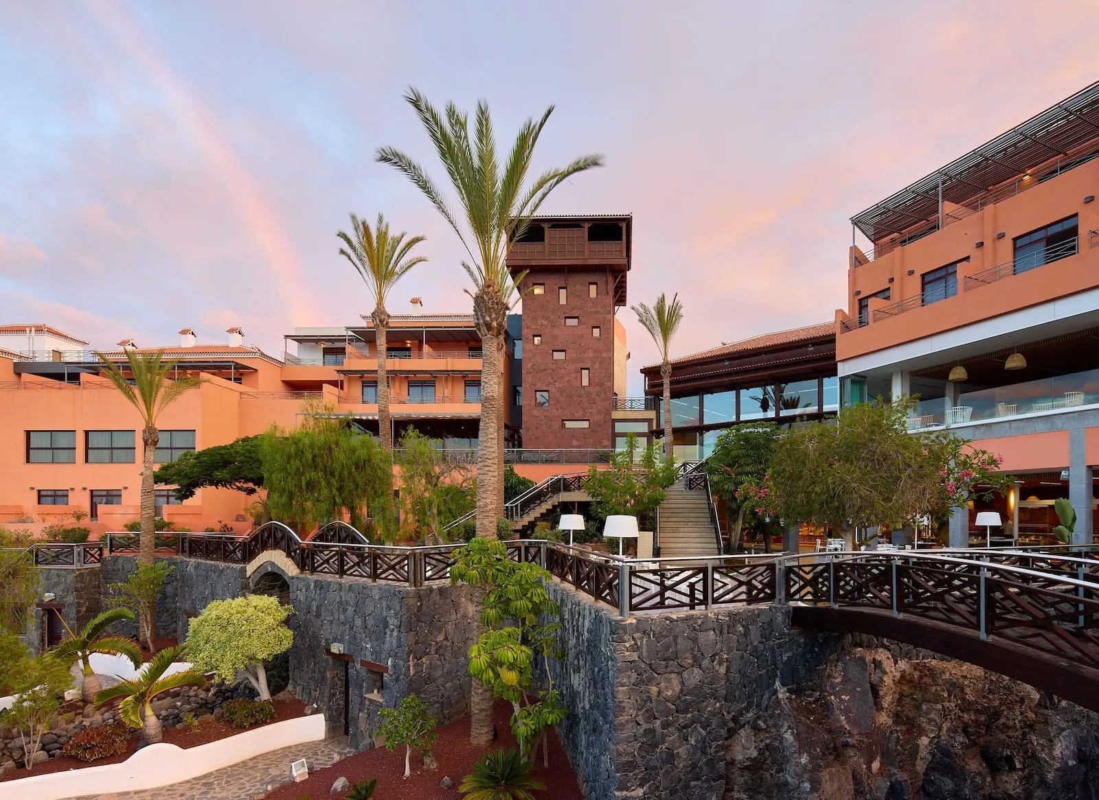 Meliá Jardines del Teide: A hotel with a stone wall, palm trees, and a staircase leading to a balcony