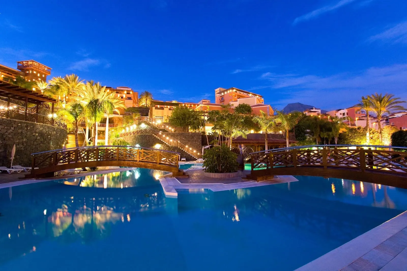 Meliá Jardines del Teide: A swimming pool with a wooden bridge, palm trees, and buildings in the background