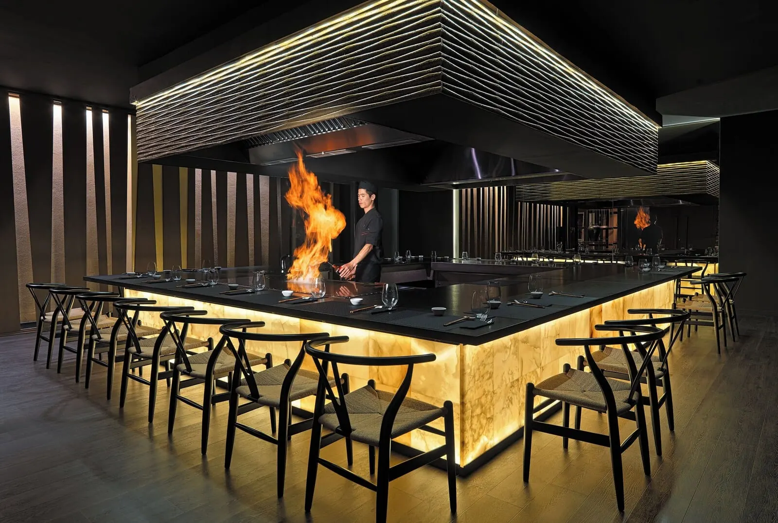 H10 Costa Adeje Palace: A modern bar area with a central island, bar stools, and a chef performing a fire act