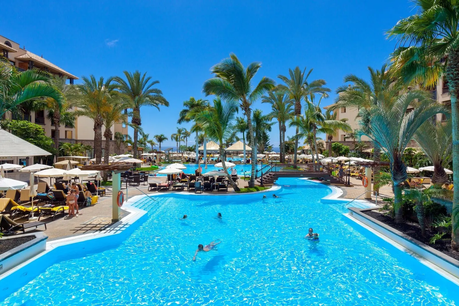 GF Gran Costa Adeje: A large pool surrounded by palm trees and lounge chairs, with a clear blue sky above