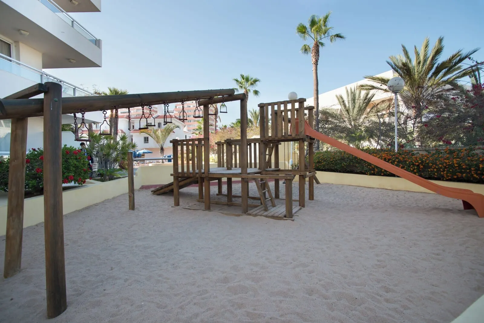 Catalonia Oro Negro: A wooden play structure in a sandy area with palm trees and a building in