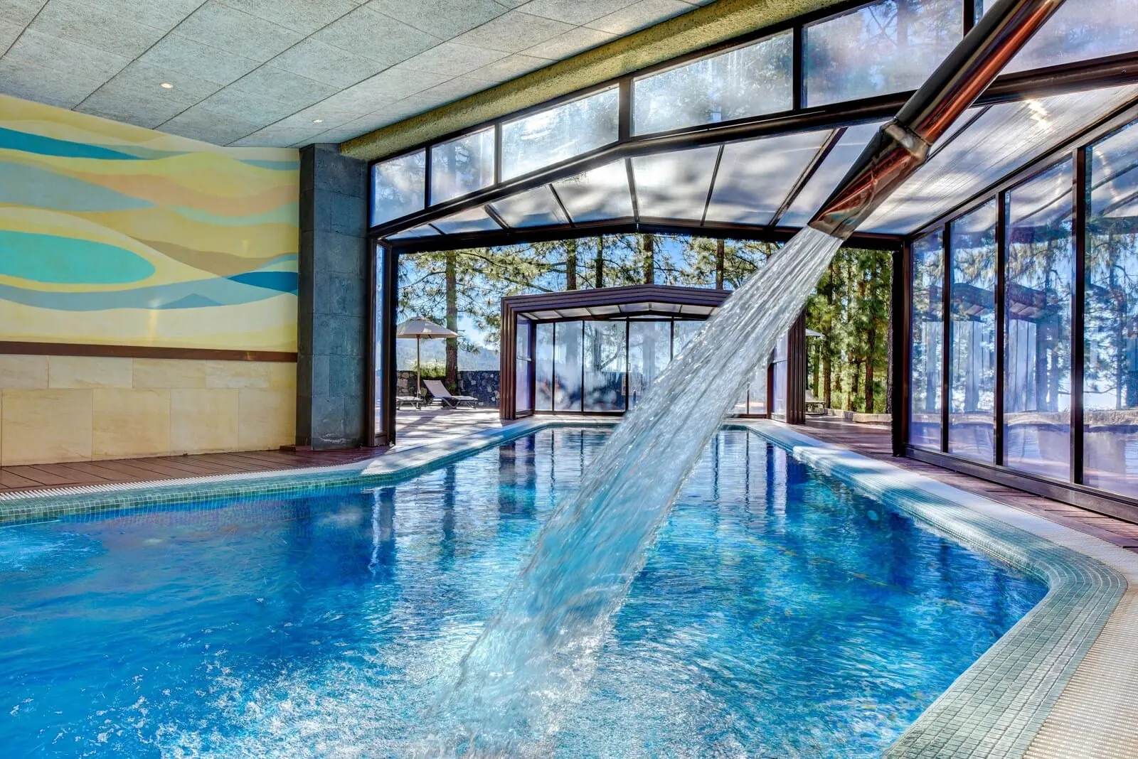 Hotel Spa Villalba: An indoor swimming pool with a water slide, large windows, and a view of trees