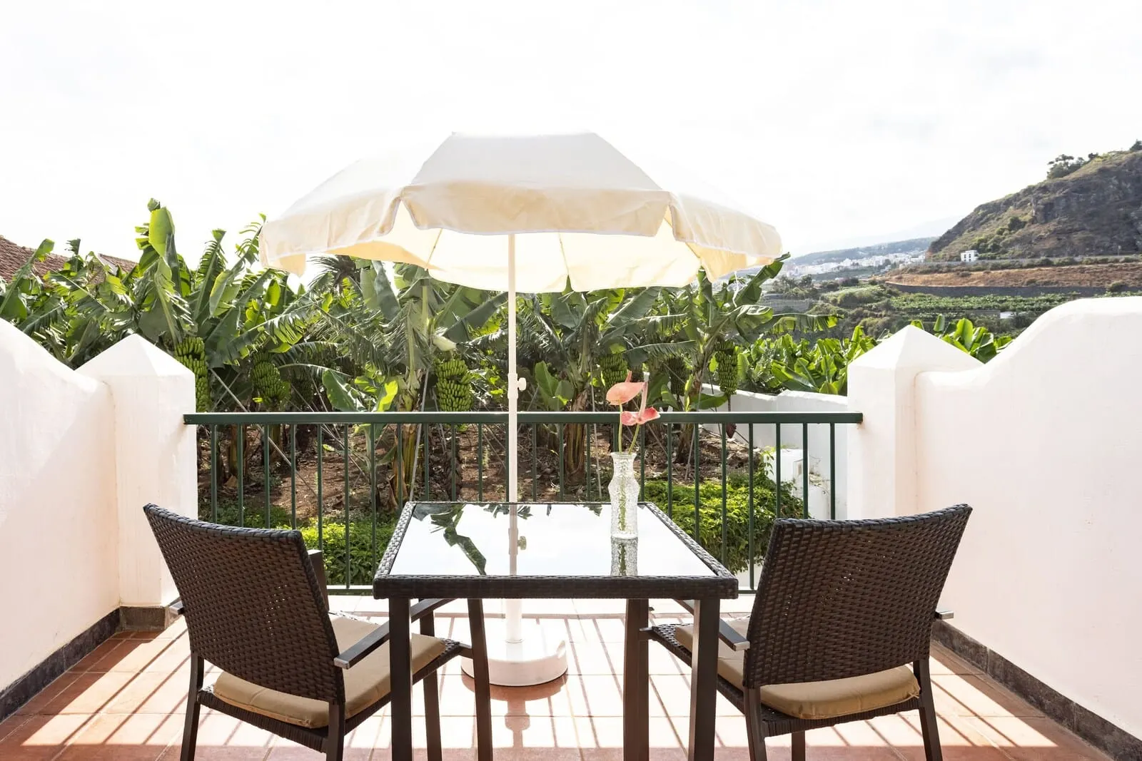 Hotel El Patio: A balcony with a table and chairs, a white umbrella, and a view of a banana plantation
