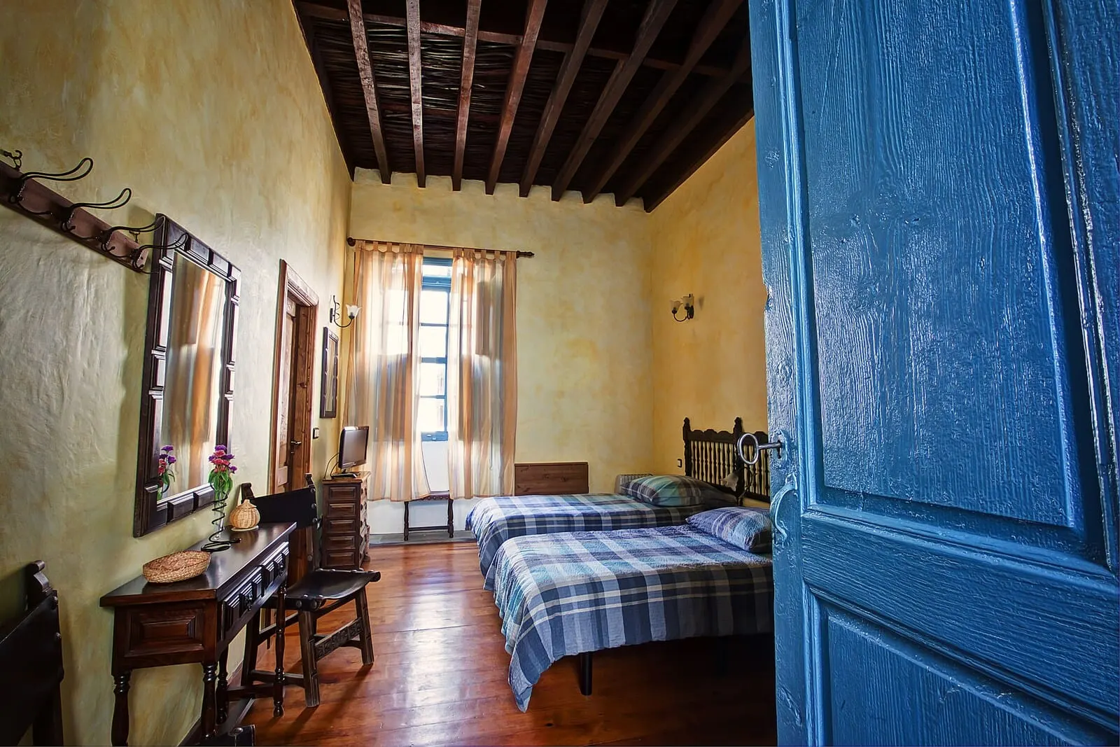 Hotel Cuatro Esquinas: A room with wooden floors, a bed with a plaid bedspread, a chair, a dresser, a mirror