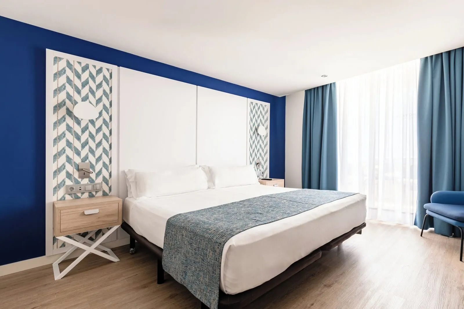 Catalonia Oro Negro: A modern hotel room with a large bed, a bedside table, a nightstand, and a chair