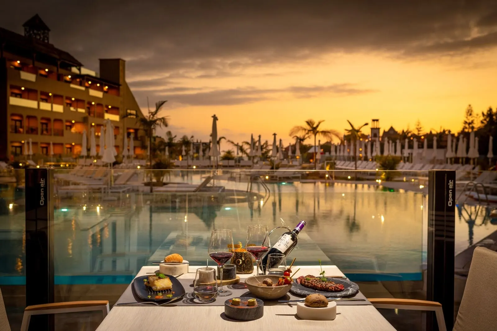 GF Victoria: A dining table with a sunset view, a pool, and a hotel building in the background