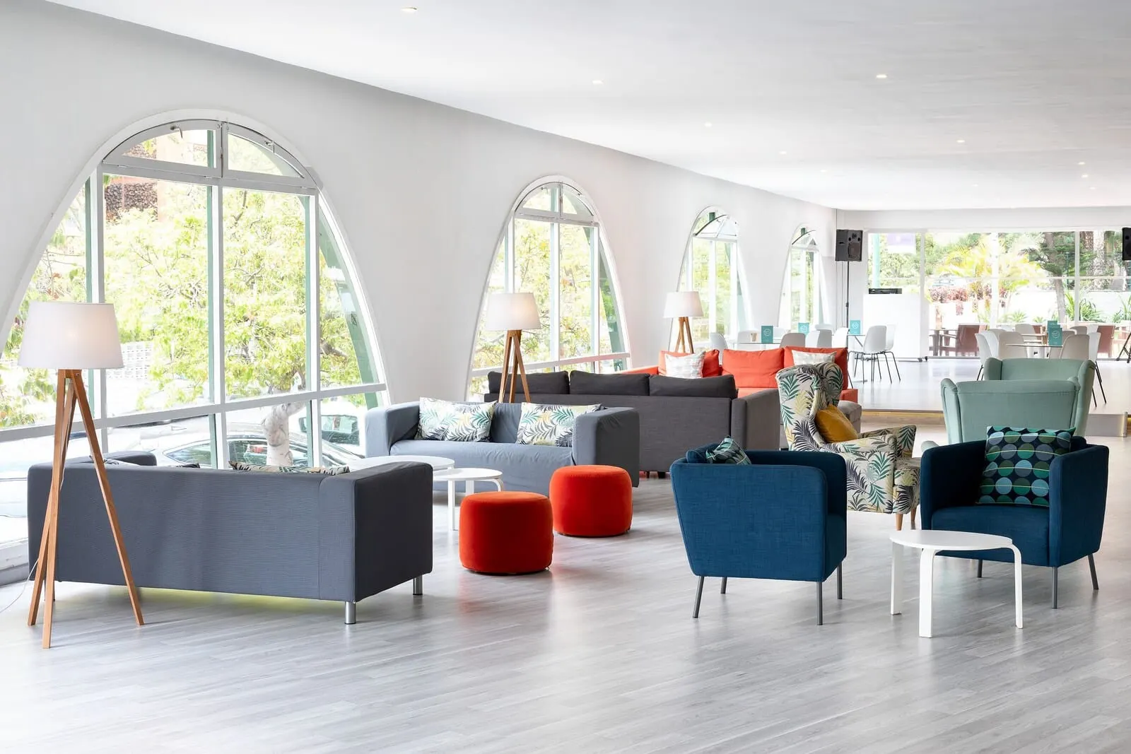 Sol Puerto de la Cruz Tenerife: A modern living room with large windows, a grey sofa, red ottomans, and blue armchairs