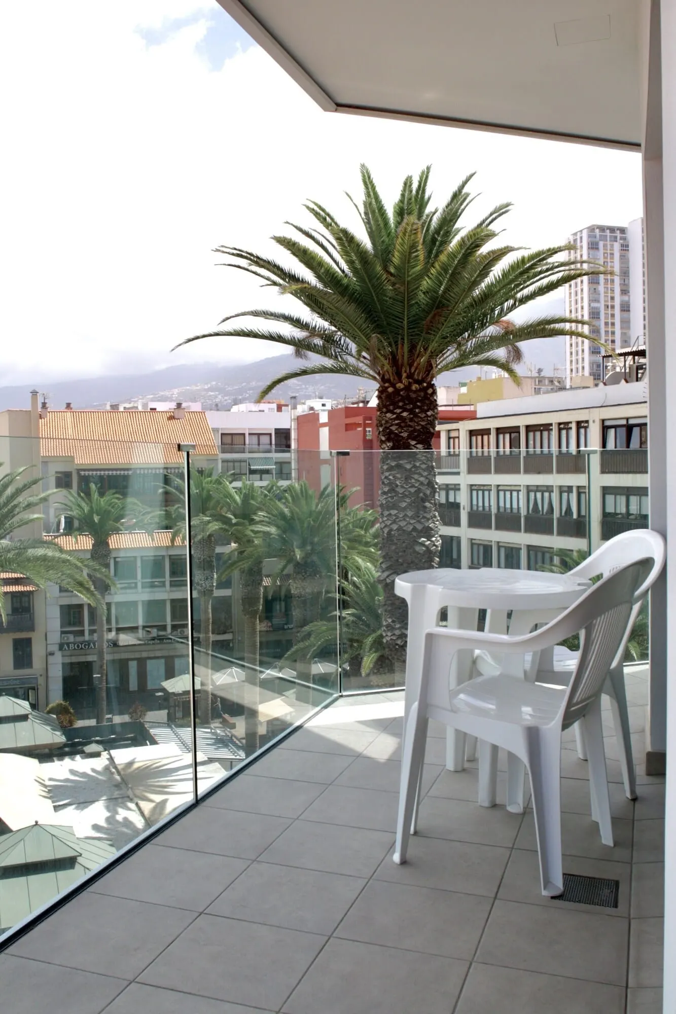 Hotel Tropical: A balcony with a glass railing, a white plastic table and chairs, a palm tree