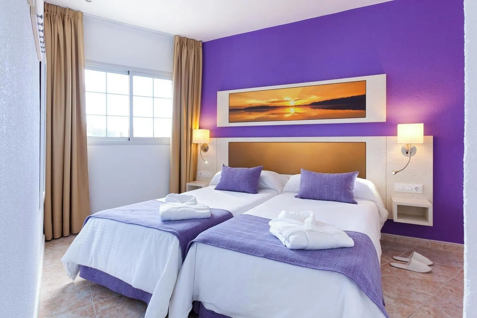 Checkin Bungalows Atlántida: A hotel room with two beds, purple walls, a sunset painting, and a window with curtains