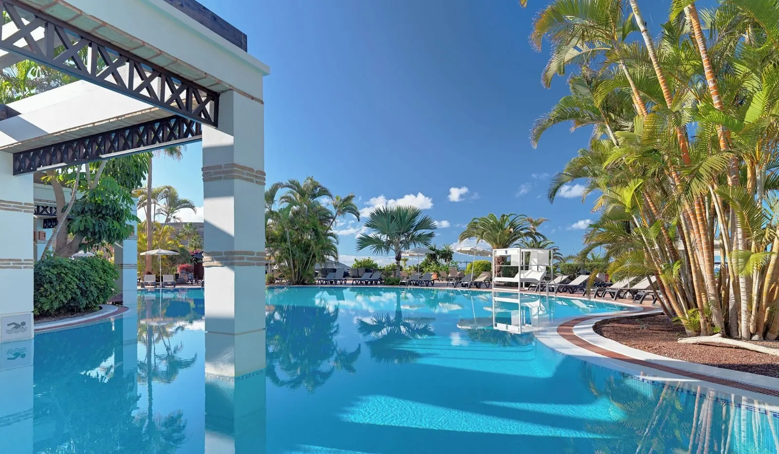 H10 Costa Adeje Palace: A clear blue pool surrounded by palm trees and lounge chairs