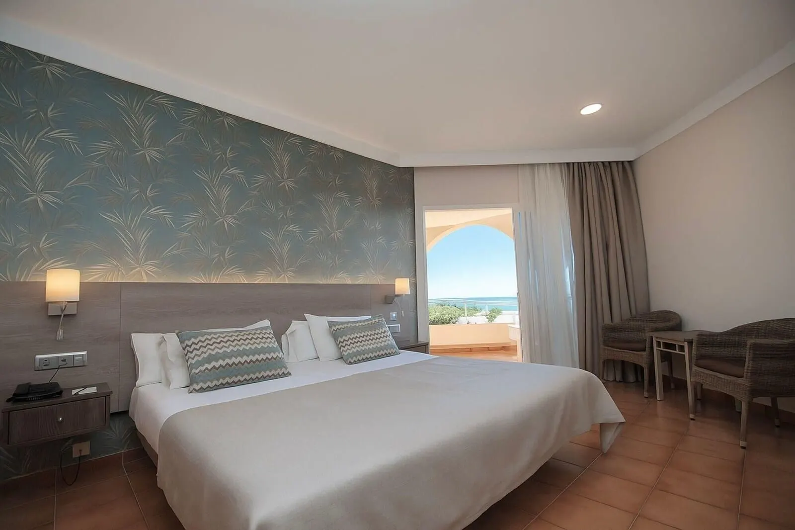Hotel Puerto Palace: A hotel room with a large bed, two bedside lamps, a nightstand, a wicker chair